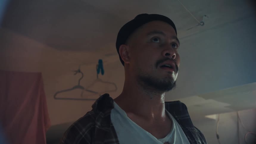 A sweaty man in a beanie gazes in awe at an unseen light source in a dimly lit basement laundry room