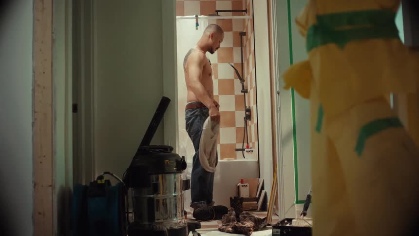 A tired handyman takes off his work clothes in a messy bathroom under renovation to take a shower after a long day