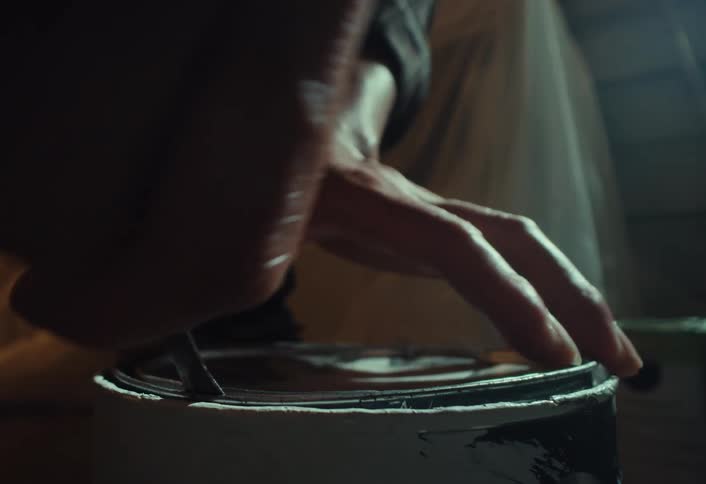 A close-up shot shows a person's hands slowly prying open a can of white paint
