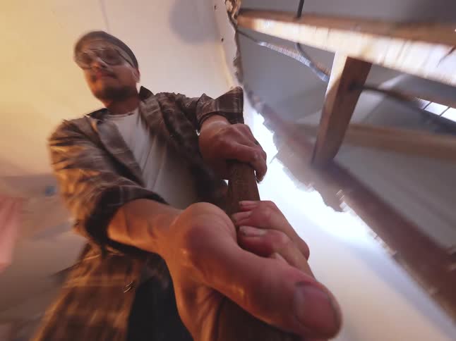 Low-angle POV shot of an aggressive man swinging a sledgehammer down to demolish a floor in a dusty, dimly lit room