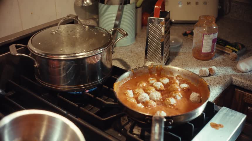 Meatballs in tomato sauce and water in a pot simmer on a messy kitchen stovetop