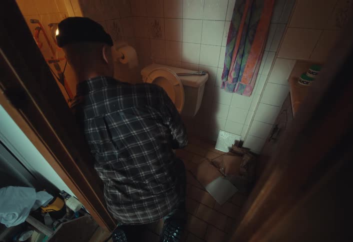 From a high angle, a sick man in a plaid shirt and hat kneels over a toilet in a grimy, poorly-lit bathroom