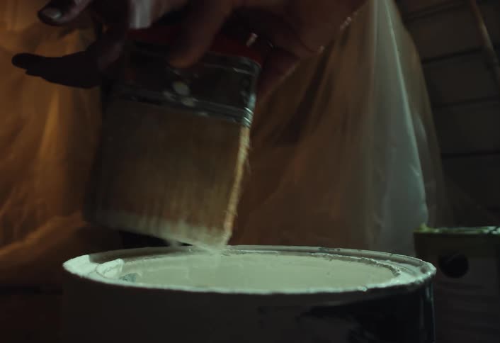 A painter dips a brush into a can of white paint and lets the excess drip off in dramatic slow motion
