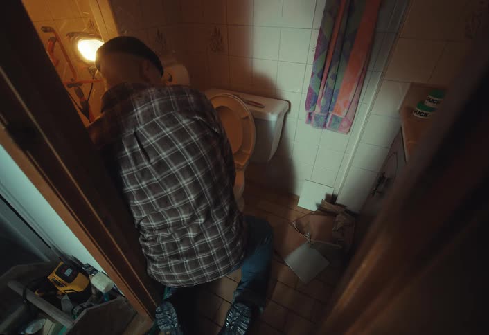 High angle shot of a sick man in a plaid shirt vomiting into a toilet in a dimly lit, messy bathroom