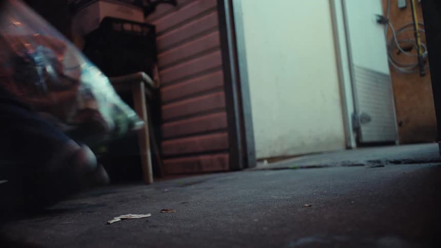 A low-angle shot captures a person dragging a large bag of trash out of a back door at night