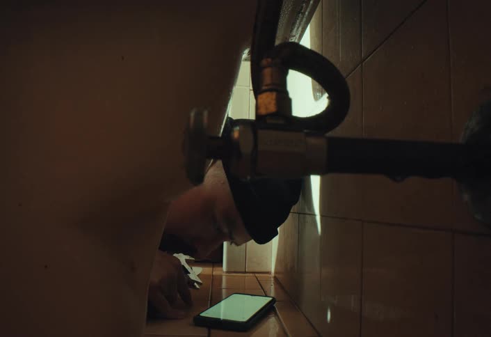 A male plumber lies on the floor under a sink watching a tutorial on his phone to fix a leaking pipe in a dimly lit bathroom