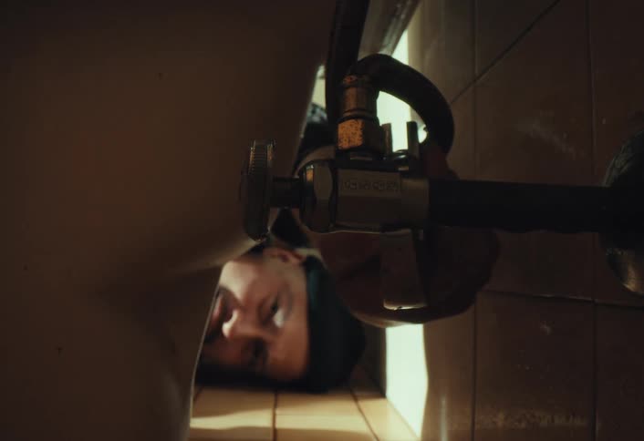 A tense man lies on a grimy tiled floor as water drips onto his face from a leaking pipe in a dimly lit room