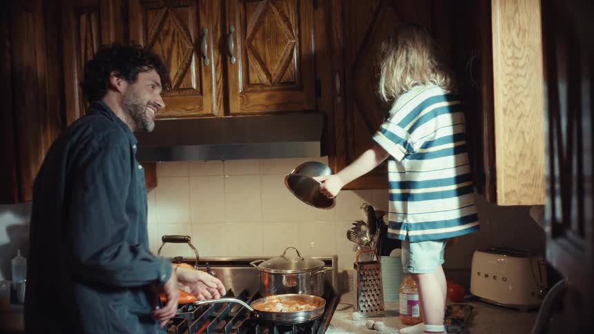 A happy father and his young son laugh and bond while cooking a messy meal together in their warm kitchen in slow motion