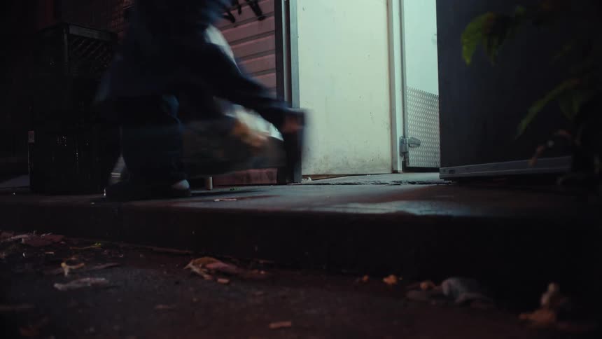 A low-angle shot of a person dragging a garbage bag out of a back door into a gritty urban alley at night