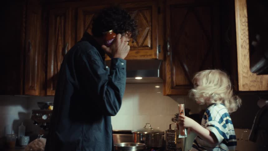 A father playfully pretends to talk on a pasta sauce jar 'phone' while cooking dinner with his laughing toddler in the kitchen