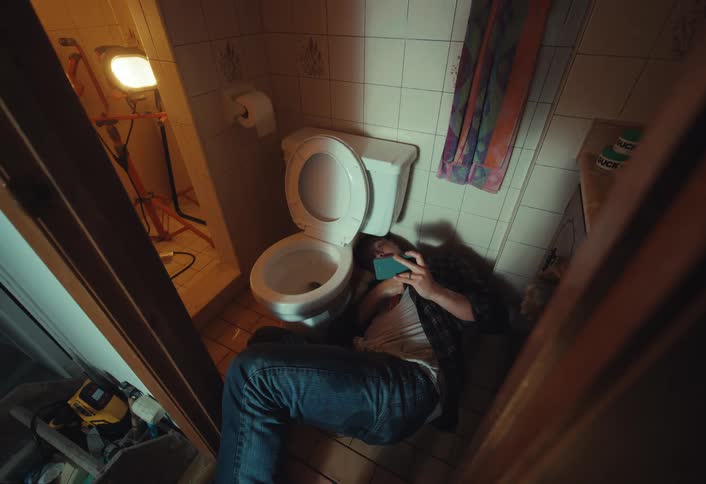 A disheveled man sits on the floor of a dimly lit, grungy bathroom, mindlessly scrolling on his smartphone