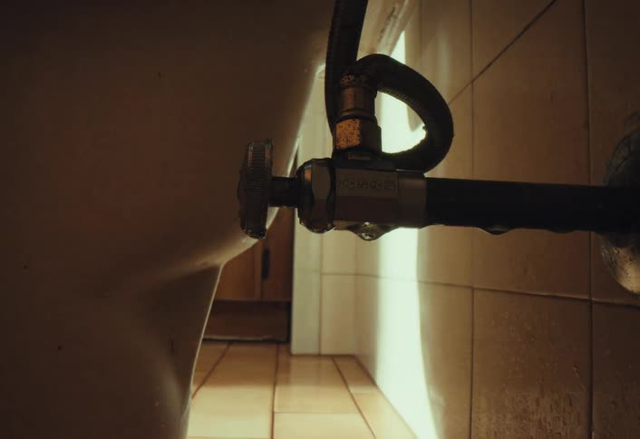 A low-angle shot of a leaky pipe dripping water behind a toilet in a dark bathroom as a mysterious shadow passes by