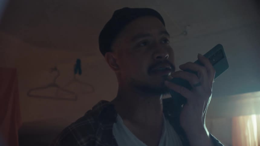 A serious young man in a beanie talks on his smartphone's speaker in a dimly lit, hazy room