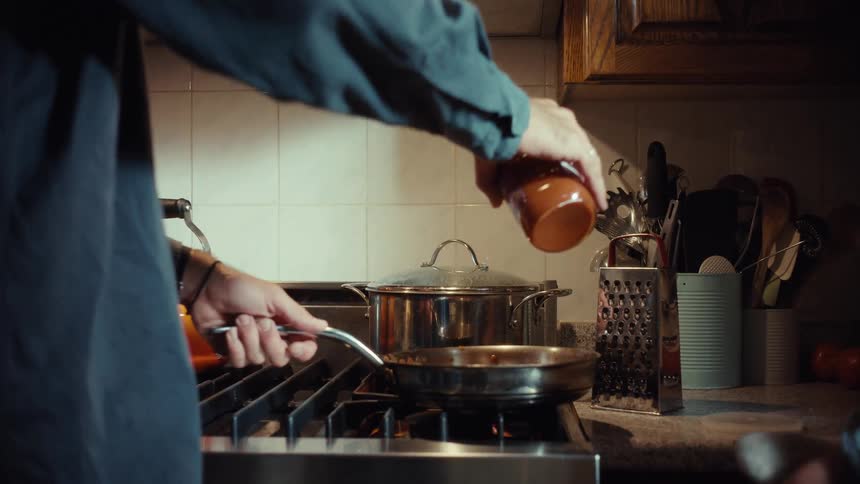 A father pours tomato sauce into a pan as his young son sits on the counter and watches him cook dinner