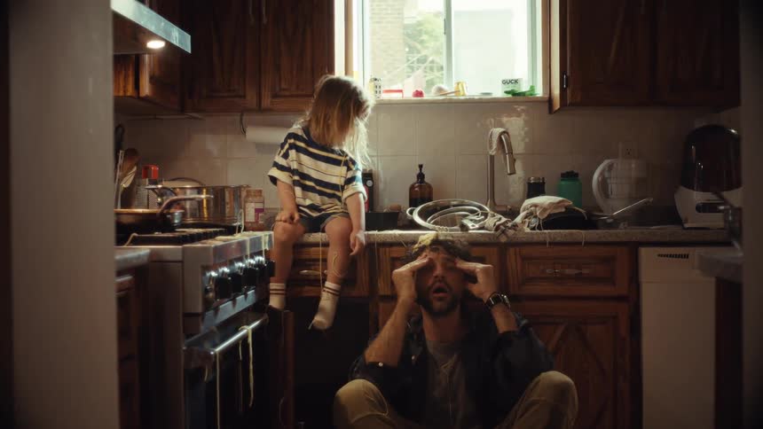 A tired father, overwhelmed in a messy kitchen, finds a moment of playful connection with his young son