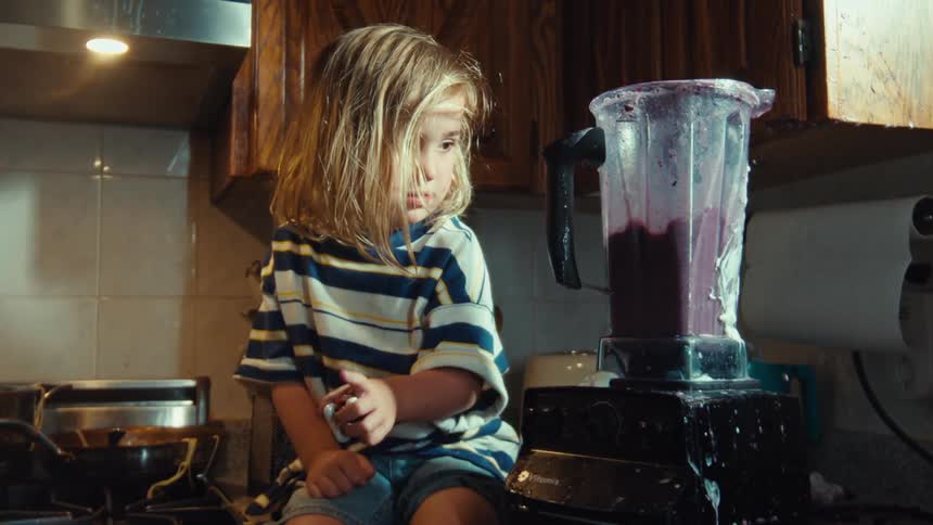 A curious young child watches intently in slow motion as a purple smoothie splashes messily from an overflowing blender in the kitchen