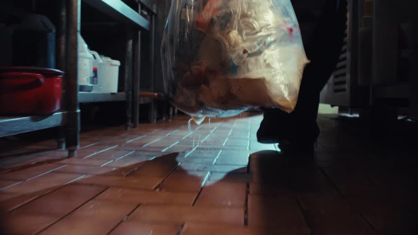 A person drags a heavy, leaking trash bag across a dirty kitchen floor in dramatic slow motion