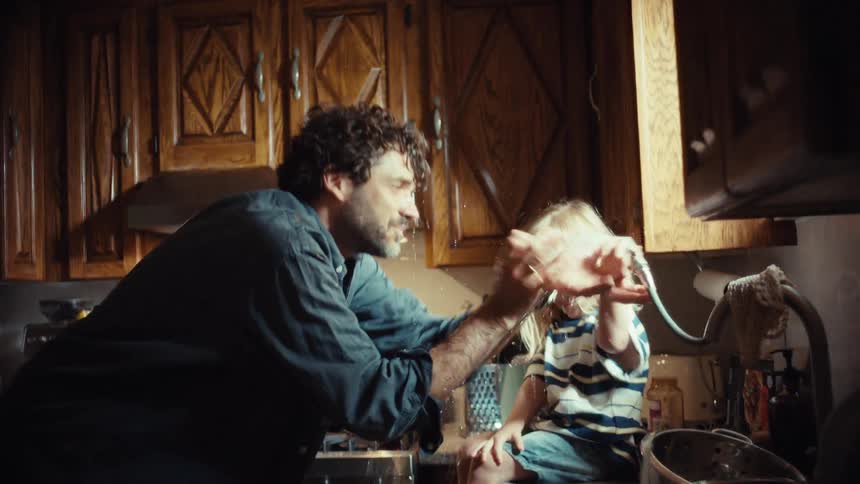 A cheerful father and his adorable daughter share a playful moment, splashing water from the kitchen sink hose