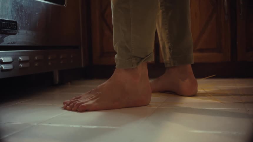 A barefoot person clumsily drops spaghetti and meatballs, creating a mess on the kitchen floor in slow motion