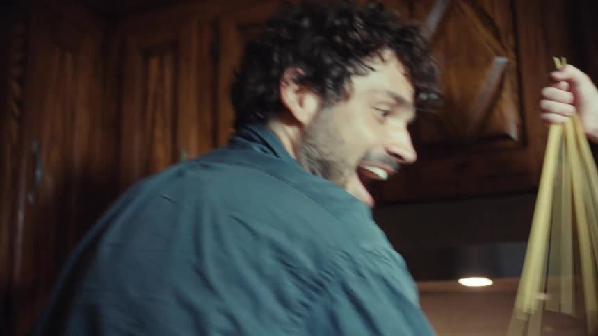 In slow motion, a smiling child playfully pulls spaghetti noodles from a package while his father cooks meatballs in a pan on the stove