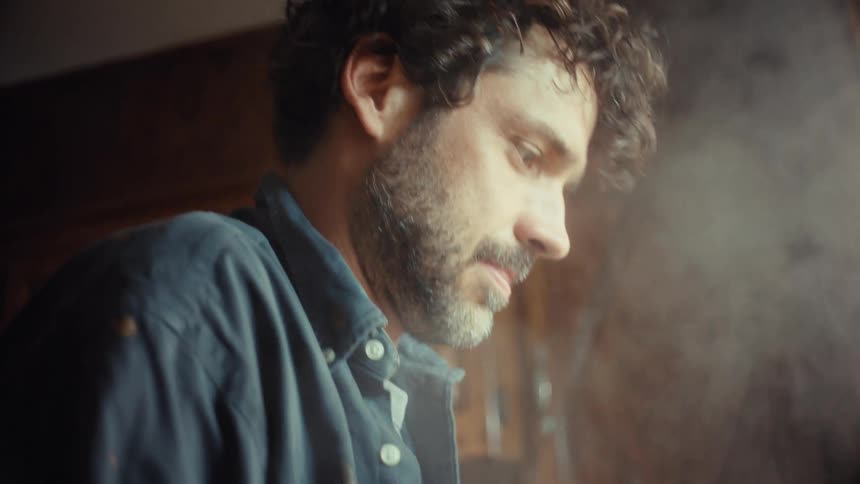 A handsome man with dark curly hair and a beard cooks in a steamy, sunlit kitchen in slow motion