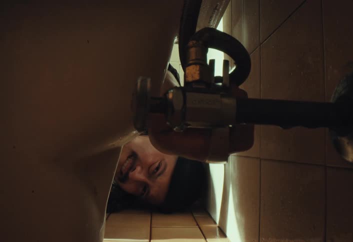 A male plumber in a beanie lies on a tiled bathroom floor, struggling to fix a leaking toilet pipe with a wrench