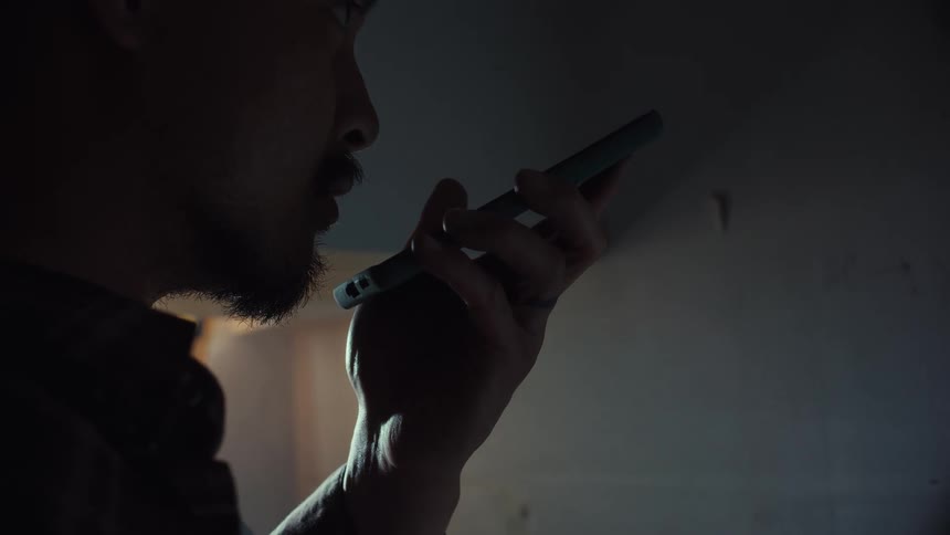 A close-up shot of a man speaking into his smartphone on speaker in a dark and moody room