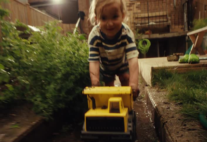 A joyful toddler covered in mud smiles while pushing a big yellow toy dump truck through a muddy path in the backyard