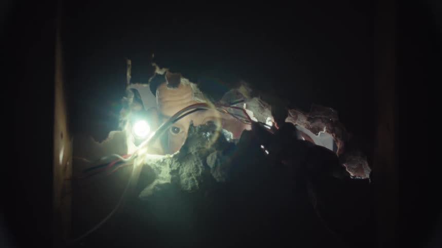 View from inside a wall as a person with a headlamp and flashlight inspects the electrical wires through a hole