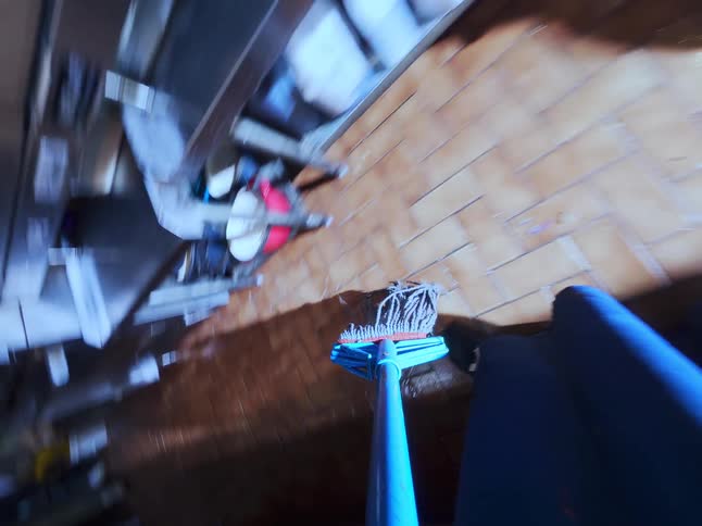 A dynamic first-person point-of-view of mopping a commercial kitchen's tiled floor with a yellow bucket