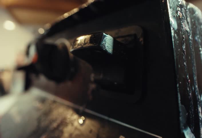 A close-up of a finger flipping the power switch on a well-used black espresso machine, releasing a puff of steam