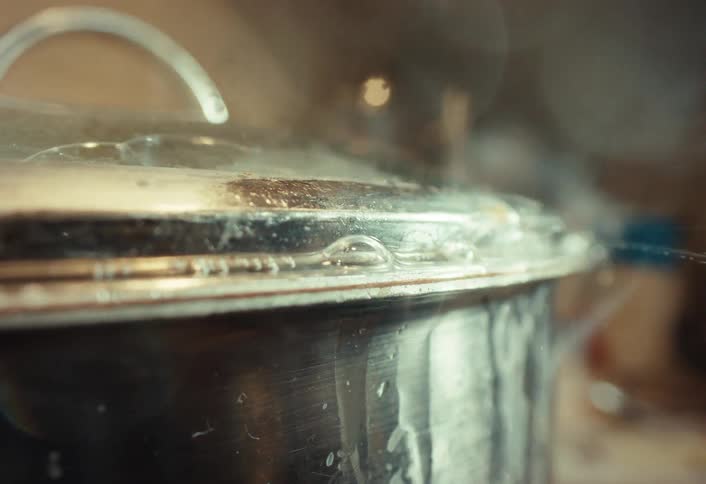 Macro slow-motion shot of a stainless steel pot boiling over, with steam and water bubbling out from under the lid