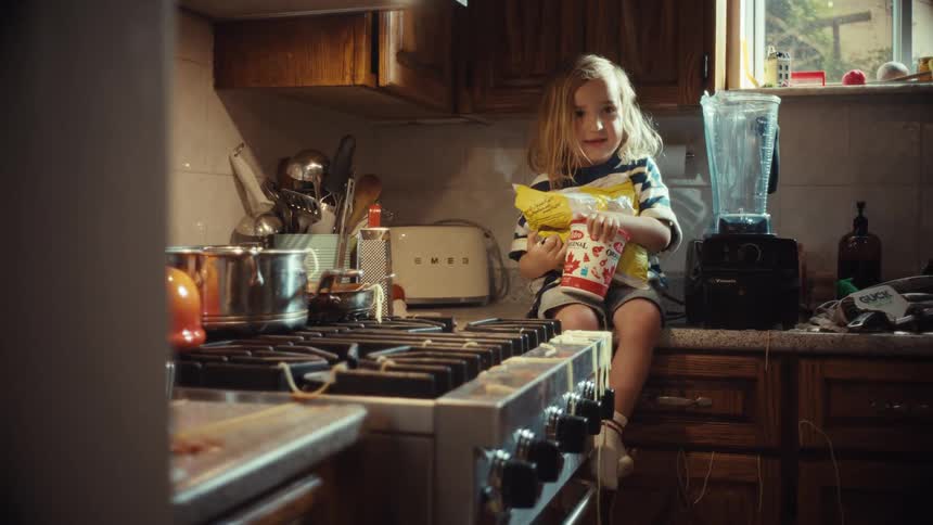 A cute child with long hair sits on a messy kitchen counter while a parent gathers ingredients for a smoothie