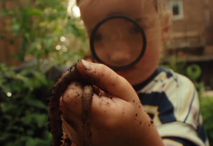 A curious young child with dirty hands holds up a long earthworm and examines it with a magnifying glass in a lush garden