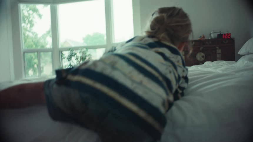 A young, muddy boy climbs up onto a clean white bed in a bedroom, leaving dirty footprints behind