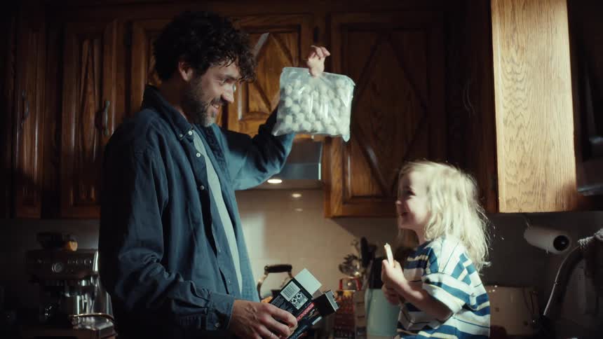 A playful father entertains his young son with a bag of meatballs while preparing dinner together in their warm, cozy kitchen