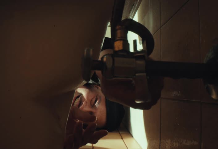 A determined man uses a wrench to tighten a pipe fitting behind a toilet in a dimly lit bathroom