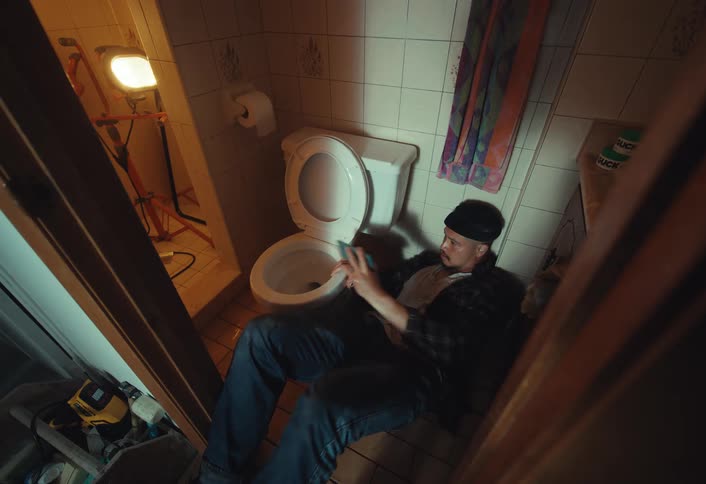 A high-angle shot captures a troubled man sitting on the floor of a messy bathroom, smoking and using his smartphone
