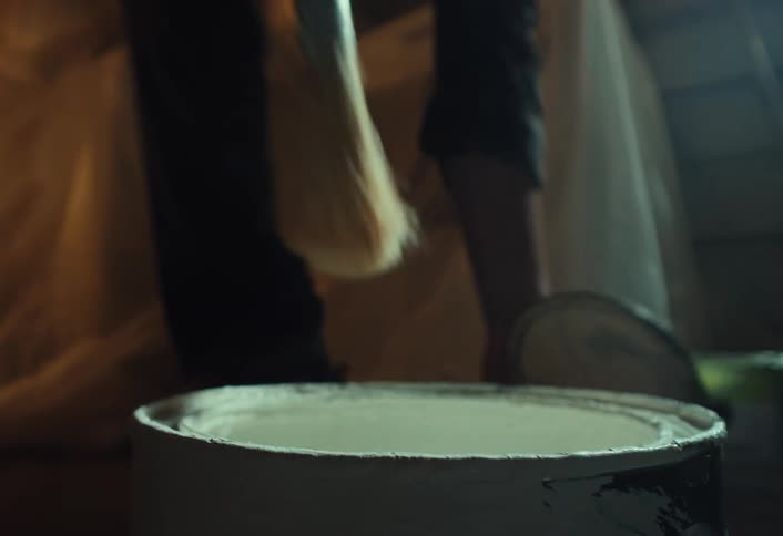 A painter carefully opens a can of white paint, stirs it with a brush, and lifts it up as the paint drips back into the can in slow motion