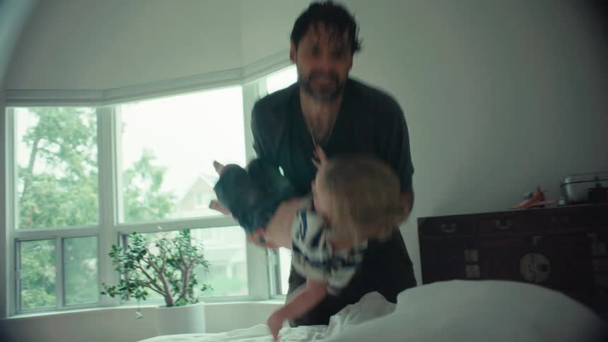 A happy father playfully swings his young son onto the bed in a bright bedroom in a dreamy slow-motion style