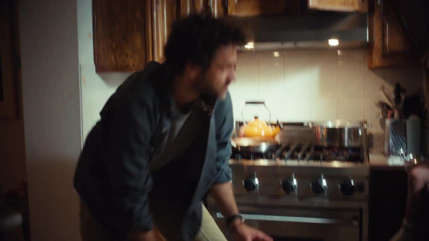 A father playfully tosses food in the air for his amused child but drops it on the floor while cooking in the kitchen