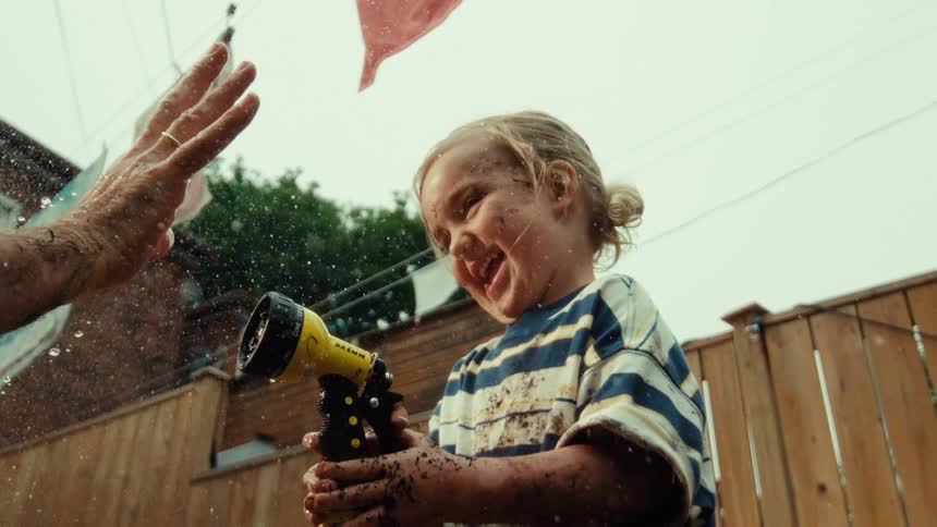 A laughing toddler covered in mud has fun spraying a garden hose in the backyard in slow motion