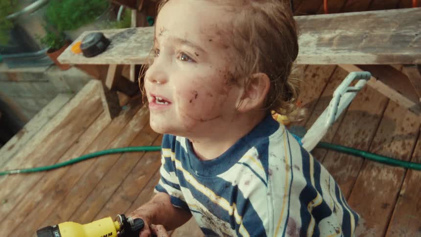 A joyful young child covered in mud smiles while spraying a water hose in the backyard in slow motion