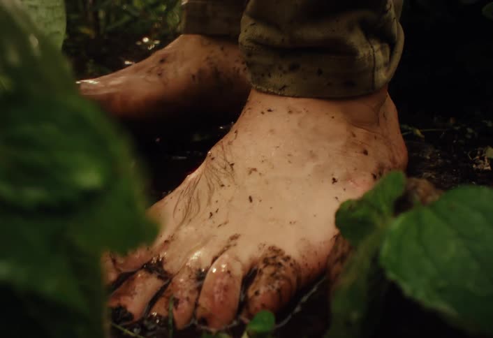 A close-up of bare feet being washed in a muddy puddle while water streams down in a lush green setting
