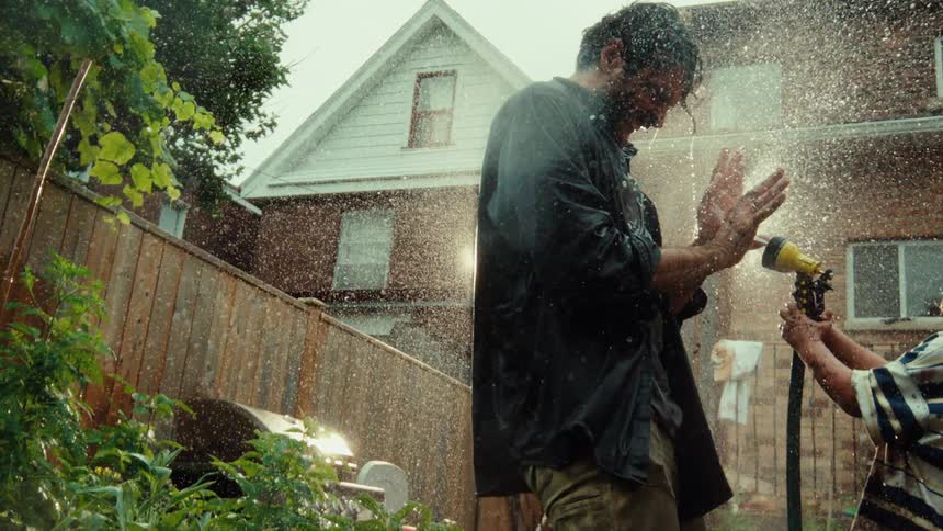 A father and his young child have a playful water fight with a garden hose in their backyard in beautiful slow motion