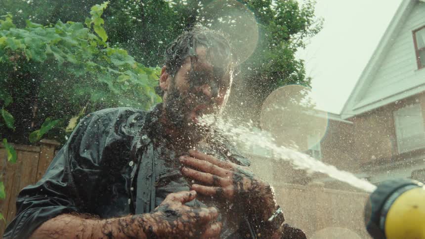 A man covered in mud is intensely sprayed with a garden hose in a suburban backyard in super slow motion