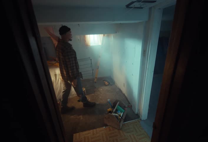 A handyman holding a hammer walks into a dusty, unfinished basement to survey the renovation project