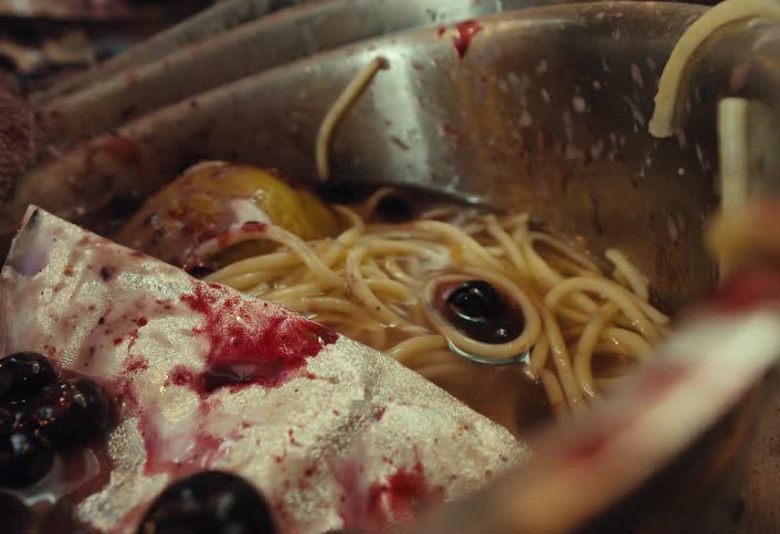 A close-up of a disgusting mix of leftover food, including a fish head and spaghetti, in a dirty bowl as more berries are thrown in