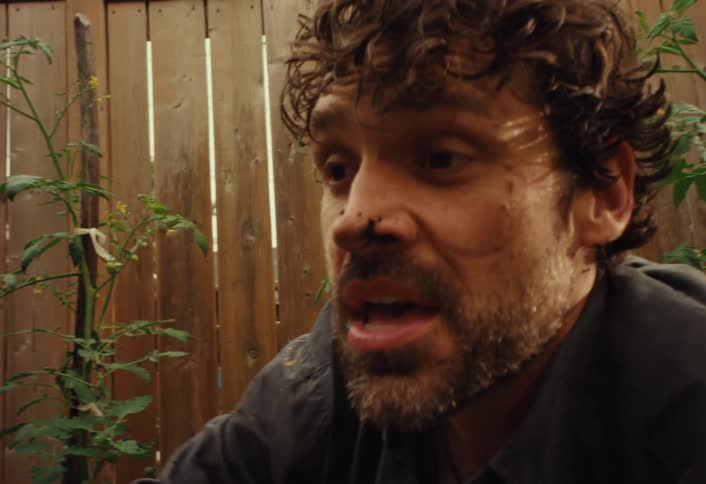 An anguished man covered in dirt expresses his sorrow and wipes his face in a garden