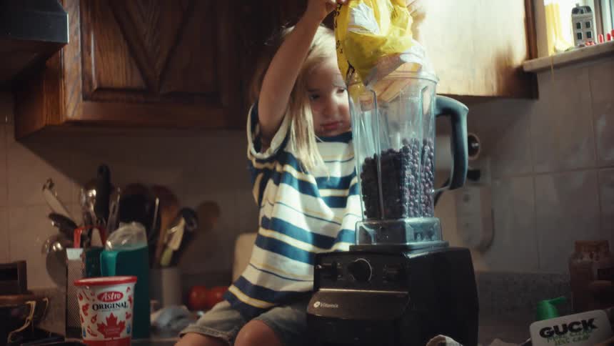 A young child sits on a kitchen counter and happily pours a bag of frozen blueberries into a blender to make a smoothie